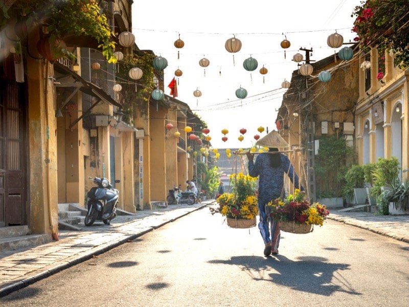 Calle del casco antiguo de Hoi An a primera hora, con faroles de seda colgados de lado a lado y una vendedora de flores cargando dos cestas con un balancín